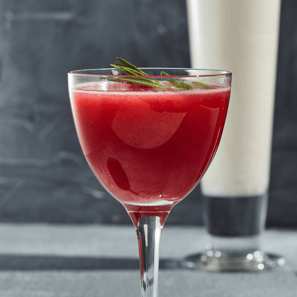 Close up of a vibrant red cocktail in a small cocktail glass garnished with Rosemary on a table.