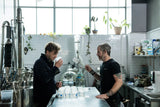 The two founders of Empirical Spirits in their distillery. They're mid-conversation across a steel workbench from each other, some distillery equipment in the background.  White subway tiles on the walls. High windows with plants on the ledge.  They appear to be sampling something as there are approximately 20 different identical containers on the table with different labels.