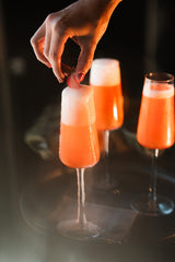 Hand placing a small glass with a red and white drink on a reflective surface with other similar glasses in the background.