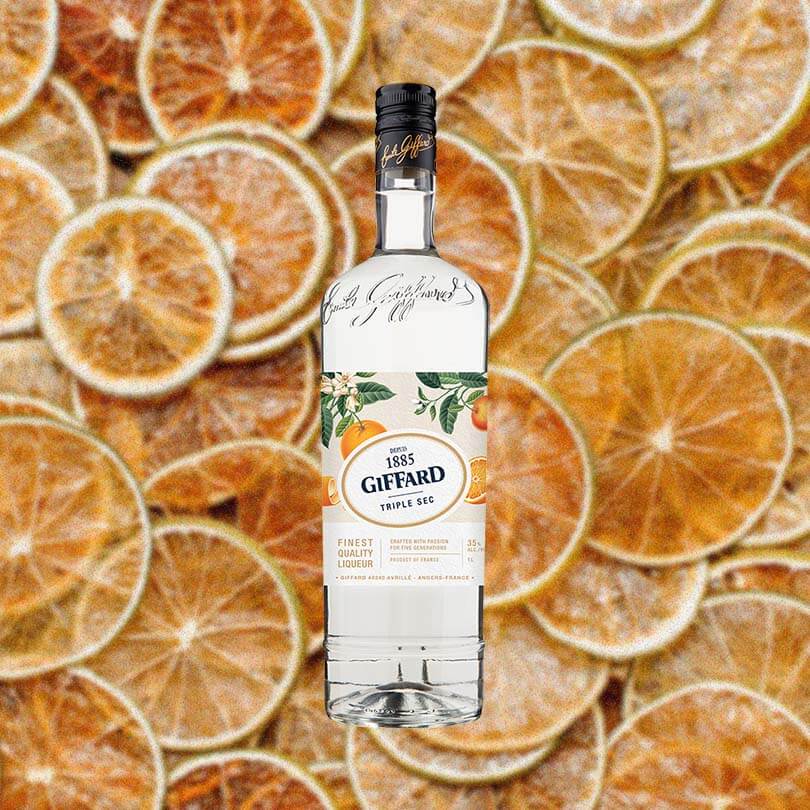A clear tall bottle of triple sec with a beautifully illustrated label featuring oranges and orange leaves. The label reads 1885 Giffard Triple Sec. This is set against a backdrop of candied orange slices artfully arranged.