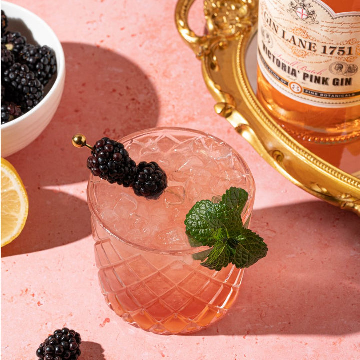 Cocktail with blackberries and mint garnish next to a bottle of Gin Lane Victoria Pink Gin on a pink surface.