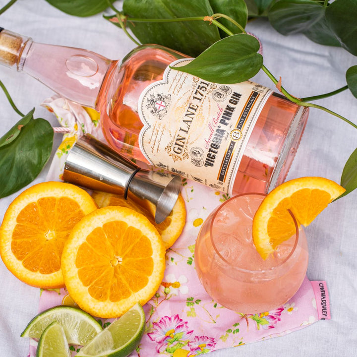 Pink gin bottle, cocktail glass with pink drink and orange slice, surrounded by oranges and limes on a floral cloth.