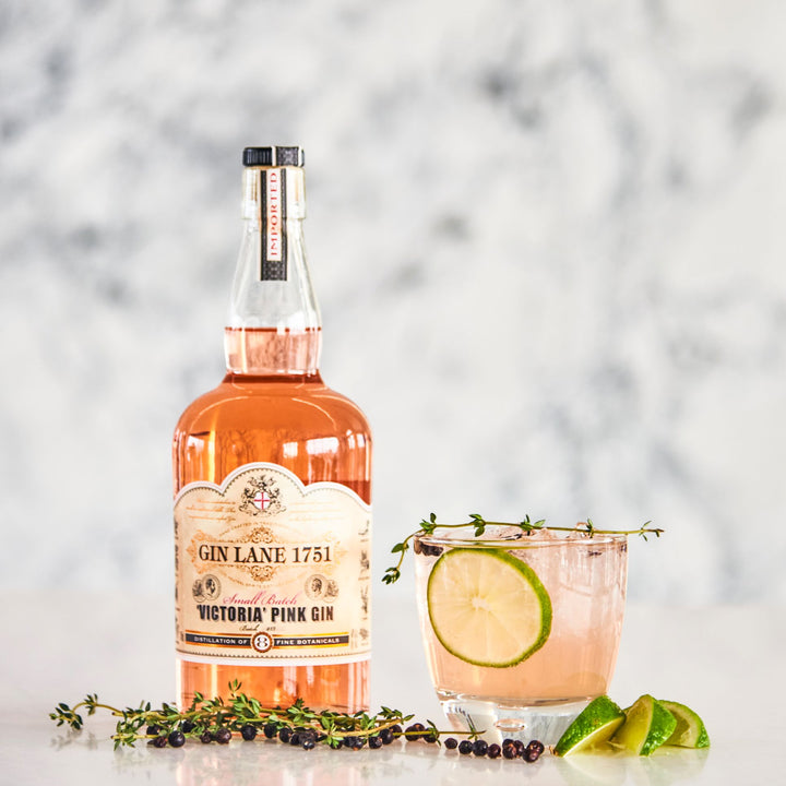 Bottle of Gin Lane 1751 Victoria Pink Gin with a cocktail garnished with lime and thyme on a marble surface.