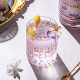 Pink cocktail with lemon and flower garnish next to a bottle of Gin Lane Violet on a white surface.