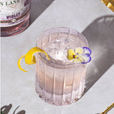 Cocktail in a clear glass with a lemon twist and flower on a light surface, with a bottle labeled 'Violet' in the background.