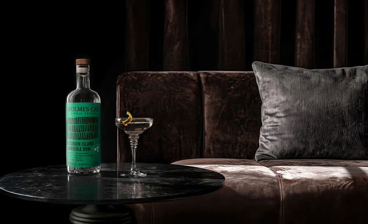 Bottle of Holmes Cay Rum and a cocktail on a marble table with a dark curtain background