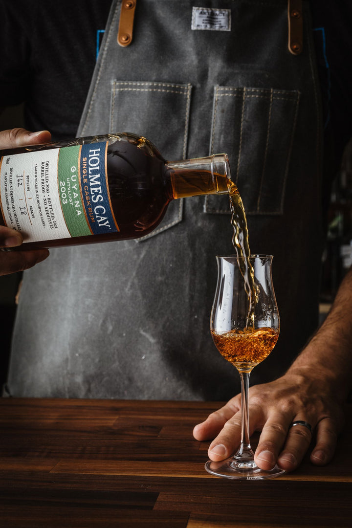 Person pouring Holman's Cay rum into a glass on a wooden surface.