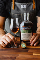 Person holding a bottle of Holmes Cay Guyana 2003 rum and a glass with rum on a wooden surface.