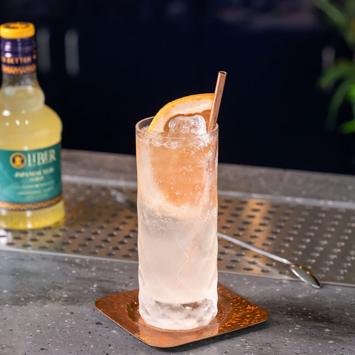 A highball glass filled with a pale pink drink, topped with a citrus slice and a metal straw, sitting on a copper coaster on a speckled gray countertop. A bottle of Liber & Co. Syrup is blurred in the background on a metal bar surface.