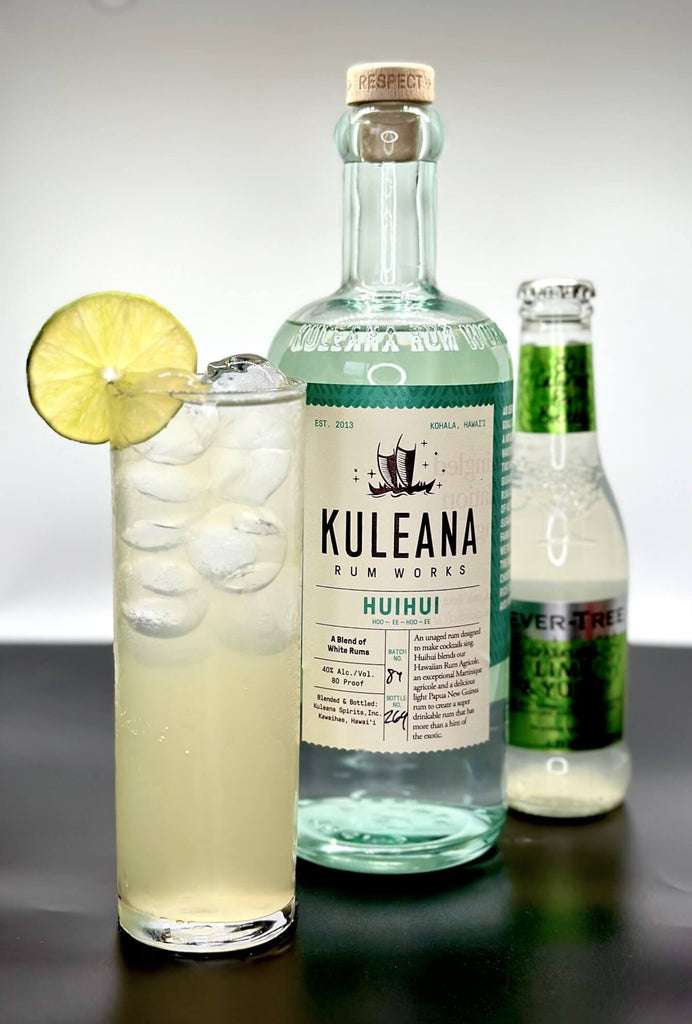 The Hawi Highball. A Collins glass with a light yellow liquid garnished with a lime wheel. In the middle a botle of Kuleana Rum Works. Just behind, a slightly out of focus bottle of Fever-Tree Lime & Yuzu soda water.