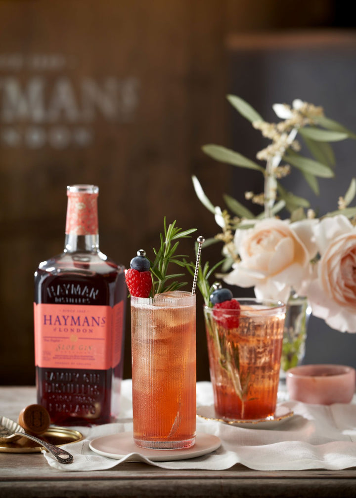 Two cocktails with a bottle of Hayman's gin on a table with flowers in the background.