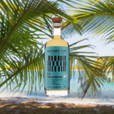 Bottle of Holmes Cay Fiji Single Origin Edition Rum over backdrop of palm trees and a beach.