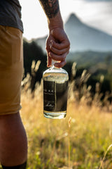A bottle of Trakal Patagonia Spirit in the foregound, a mountain in the distance. The bottle is held by a man with a tattooed arm -- you just see his partial profile from the waist down.
