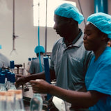 Two workers in a factory setting, wearing blue hairnets and gloves, handling bottles.