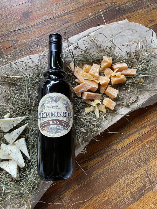 Bottle of Alpeggio Hay Liqueur on a wooden surface with hay and cheese.