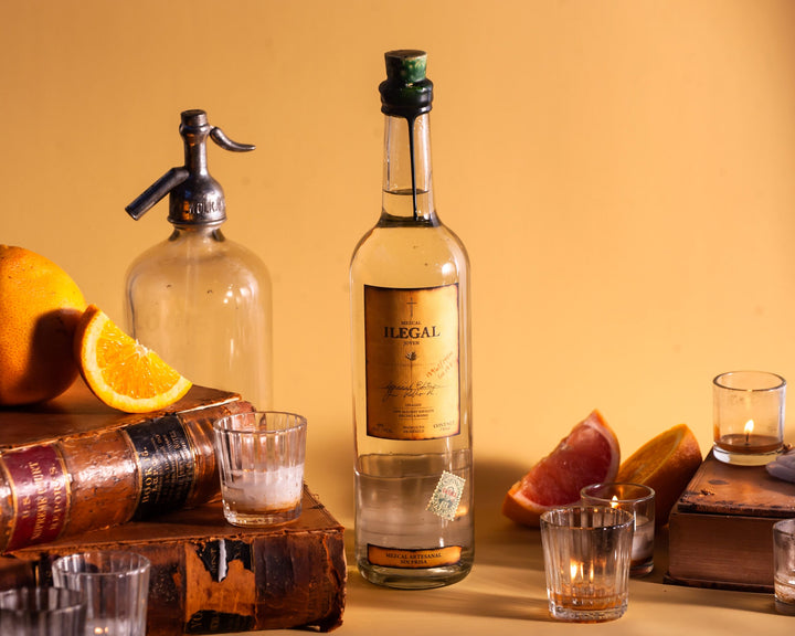 Bottle of Illegal tequila with oranges, a seltzer bottle, and shot glasses on a wooden surface.