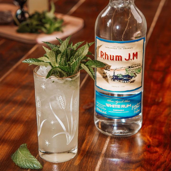 Rum cocktail with mint leaves and a bottle of Rhum J.M. on a wooden table.
