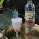 Cocktail in a glass with a garnish next to a bottle of Rhum J.M. rum on a reflective surface.