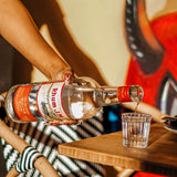 Person pouring rum into a glass on a wooden table with a colorful background