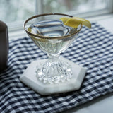 Clear glass with a lemon wedge on a marble coaster on a checkered tablecloth.