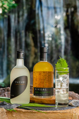 A bottle of Yuzuri Yuzu Liqueur to the left, a bottle of Kikori whiskey in the middle, and a Collins glass filled with an appealing clear cocktail garnished with a green leaf. A waterfall is in the distance behind the three bottles.