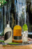 A bottle of Yuzuri Yuzu Liqueur to the left, a bottle of Kikori whiskey in the middle, and a Collins glass filled with an appealing clear cocktail garnished with a green leaf. A waterfall is in the distance behind the three bottles.