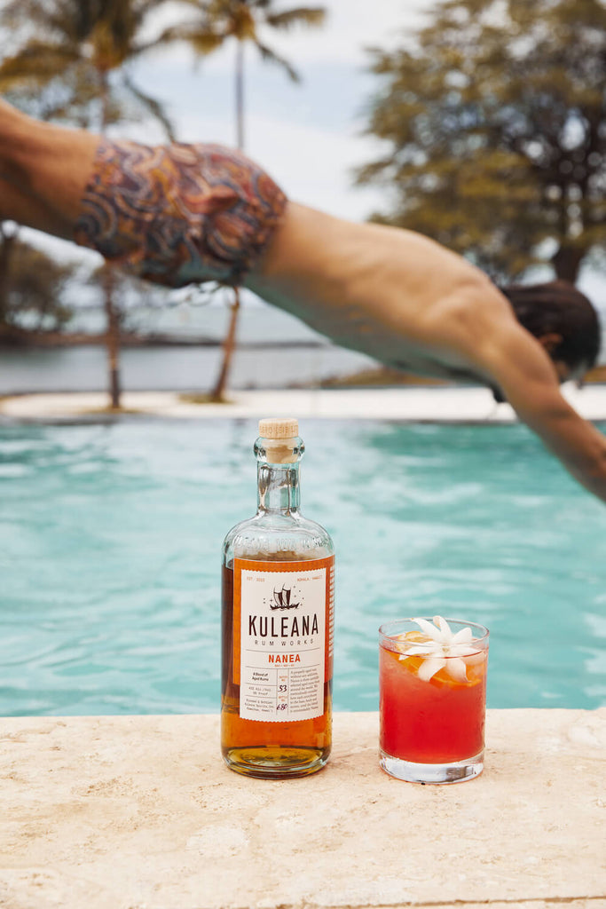 A bottle of Kuleana Rum Works Nanea Rum and a brightly colored cocktail with jasmine flower garnish in the foreground. In the background a man is diving into a pool, palm trees in the distance.
