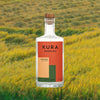 Kura Shochu bottle with a colorful label against a blurred background of rice fields in a vibrant yellow green color
