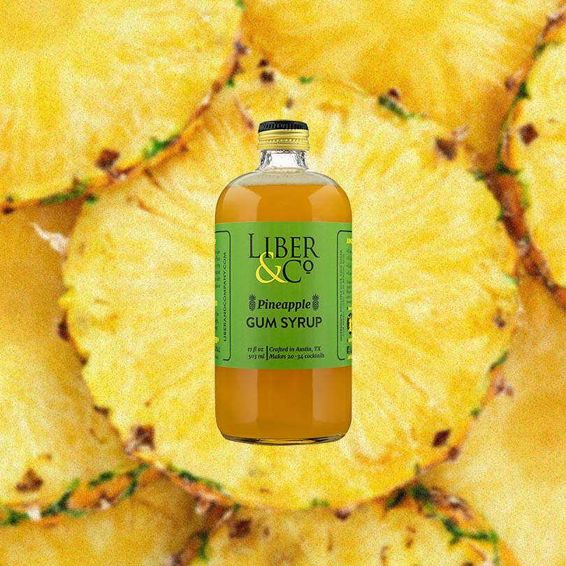 Bottle of Liber & Co. Pineapple Gum Syrup over backdrop image of sliced Pineapples.