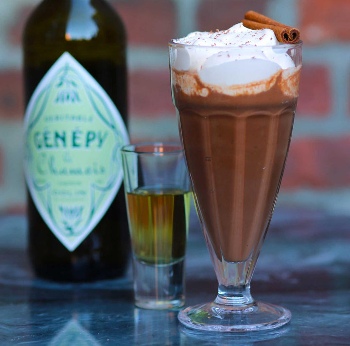 Chocolate drink with whipped cream and a cinnamon stick next to a bottle of Genever on a reflective surface.