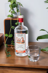 Bottle of Renegade Rum on a wooden surface with a glass of water and plants in the background.