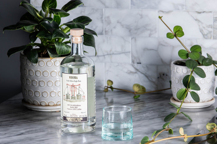 Mhoba Rum bottle with a glass on a marble surface with plants in the background.