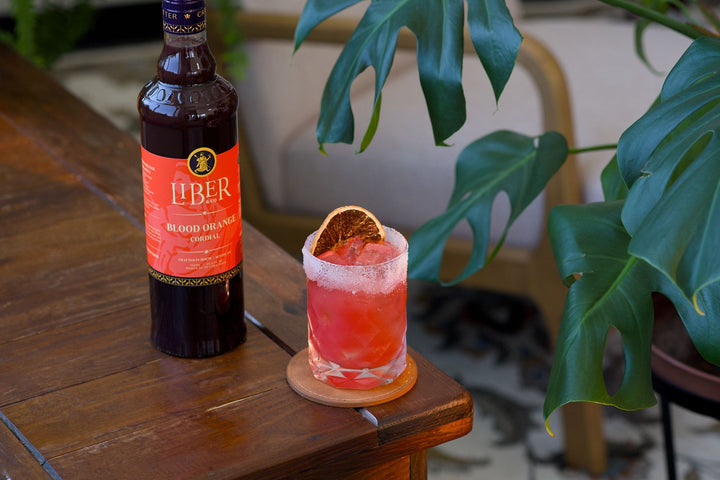 Bottle of Liber & Co Blood Orange Syrup next to a cocktail on a wooden table with plants in the background