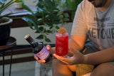 Person holding a bottle of Liber & Co and a red cocktail with a slice of orange, surrounded by plants.