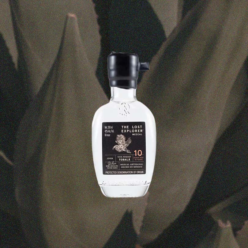 A mini sized 200ml bottle of The Lost Explorer Mezcal. The top is dipped in black wax, and the label is black with gold detailing.  It reads Mezcal Artesanal Hecho in Mexico.  10 Agave Age in Years. Against a moody macro shot of agave plants.