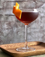 Cocktail in a textured glass with an orange twist on a wooden tray against a stone wall background