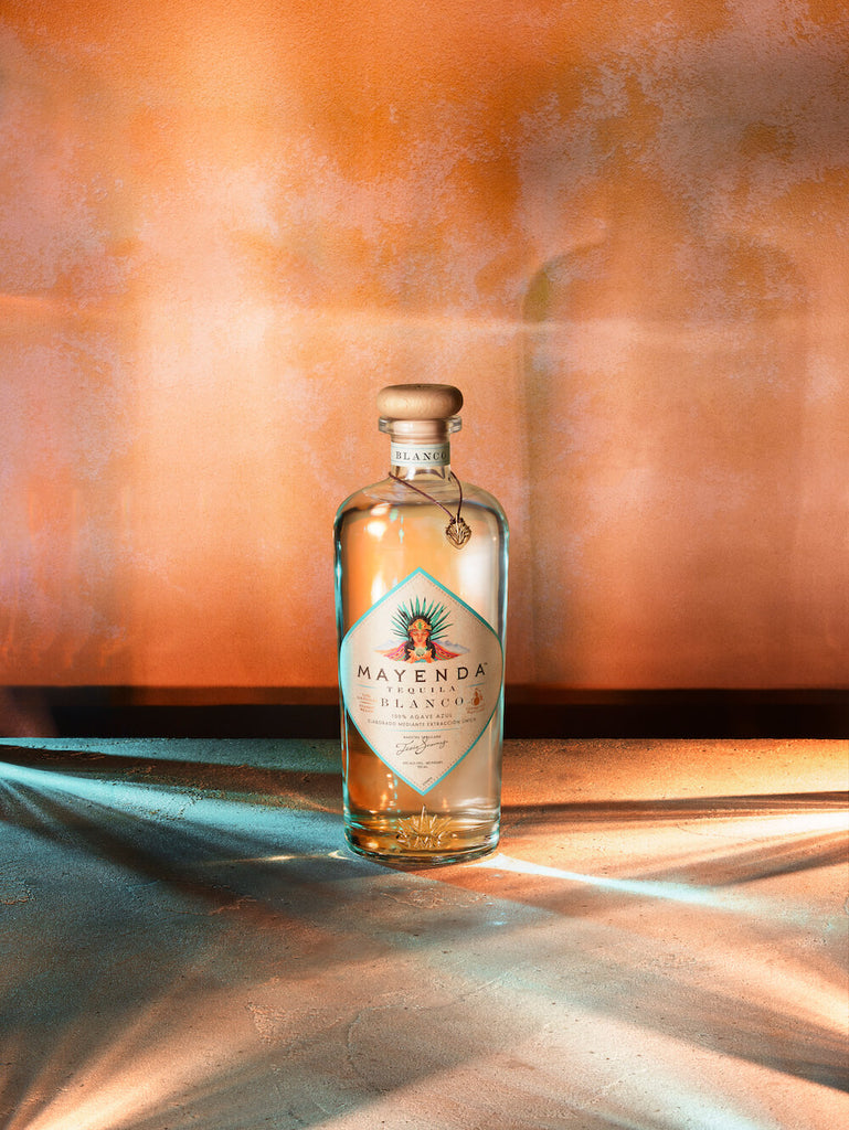A bottle of Mayenda Tequila Blanco shot against a warm red colored wall.