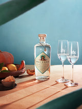 A table with a wooden bowl of squash and citrus to one side. A bottle of Mayenda Tequila Blanco sits next to it, centered in the image. To the right are two thin flutes one third full of tequila.  The backdrop is blue.