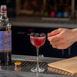 Cocktail with blackberries being garnished next to a bottle of Liber & Co Blackberry Syrup.