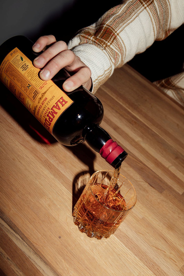 Person pouring a drink from a bottle labeled 'Hampden' into a glass on a wooden surface.