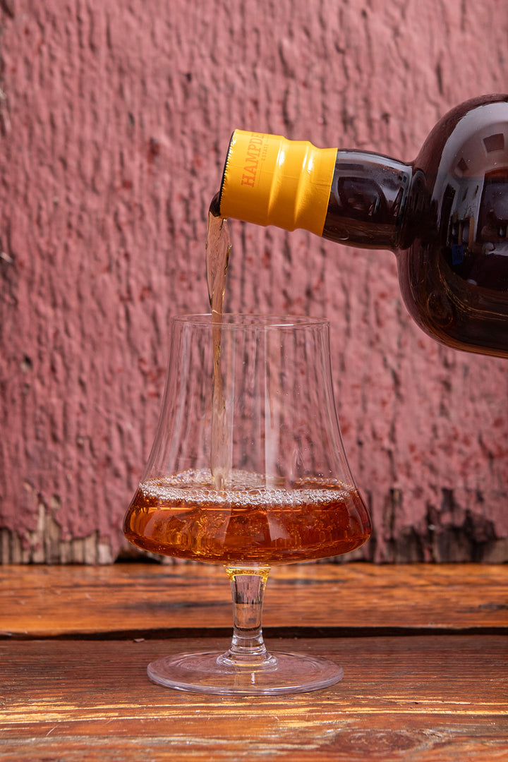 Brown liquid being poured from a bottle into a glass against a pink textured wall.