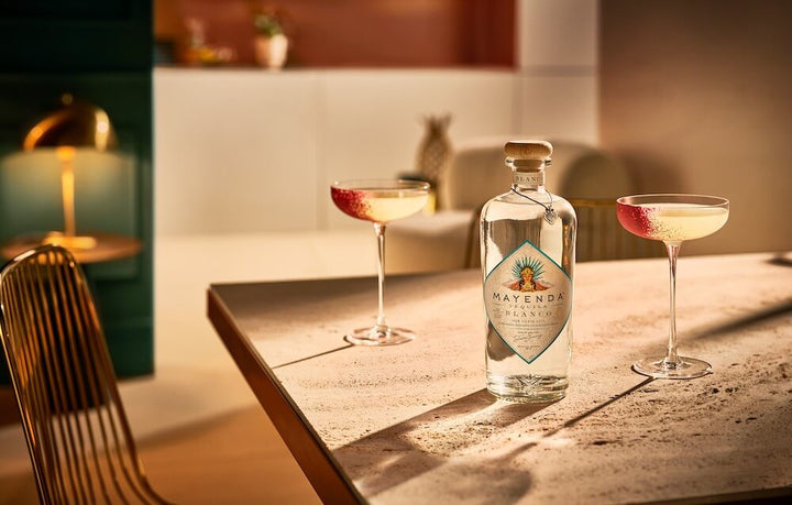 An interior shot of a kitchen. In the foreground is a bottle of Mayenda Tequila Blanco flanked on each side by an elegant, thin-stemmed cocktail glass filled with an alluring looking drink.