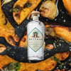 A premium bottle of clear tequila with a wood stopper. The label is diamond shaped with a teal edge, and it reads: Mayenda Tequila Blanco. 100% Agave Azul.  The backdrop is a macro shot of honey drizzled roasted squash, fragrant and delicious.