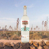 A bottle of Mezcal Union Uno set against a backdrop of flowering agave plants and a light blue sky.