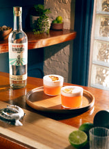 A brightly lit bar with a bottle of Mezcal Union Uno on it, sitting beside is a wooden tray with two cocktails, a pleasing orange hue, each with a light foamy top and a dehydrated orange slice garnish..