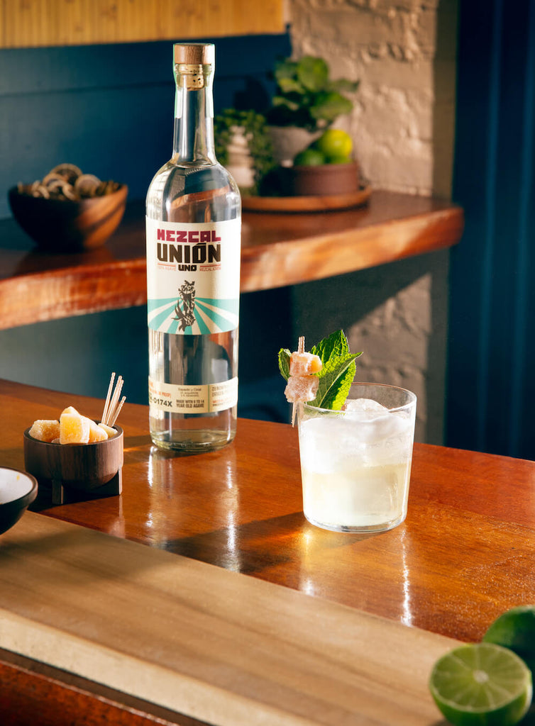 A brightly lit bar with a bottle of Mezcal Union Uno on it, sitting next to it a refreshing looking cocktail garnished with candied ginger and a mint sprig.