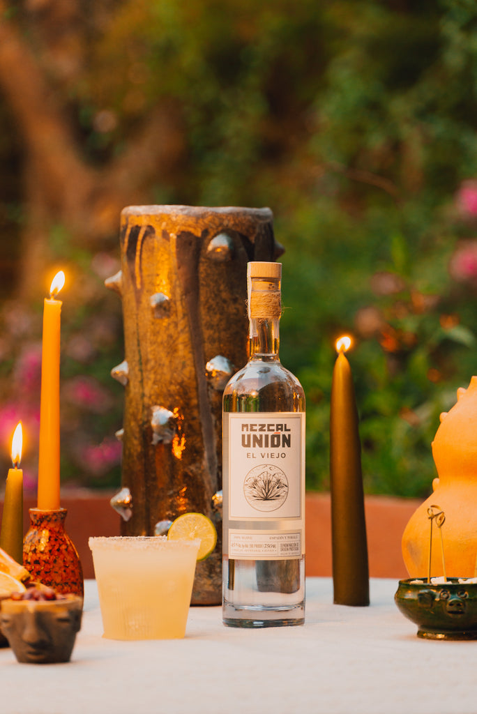 A bottle of Mezcal Union El Viejo on a beautifully set table, candles lit.  Beside the bottle is a salt-rimmed cocktail garnished with a lime wheel.