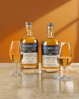 Two bottles of Holmes Cay South Africa whisky with two glasses on a wooden surface against an orange background.