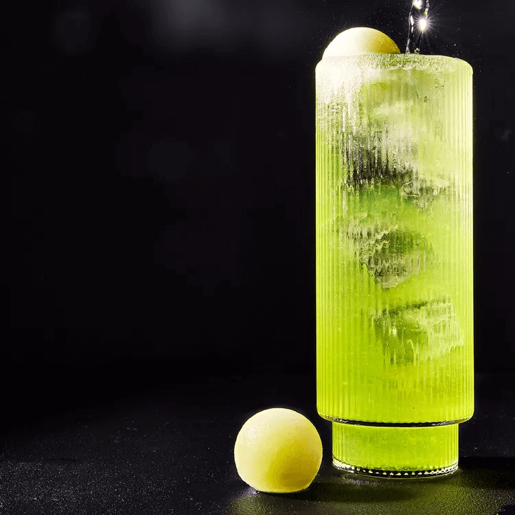 A tall art deco style cocktail glass with green liquid and ice cubs in it. A melon ball adorns the top and a spherical melon ball sits beside it.