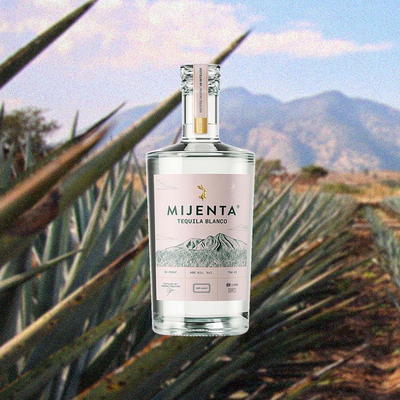 Bottle of Mijenta Blanco Tequila over backdrop of agave and a mountain side.